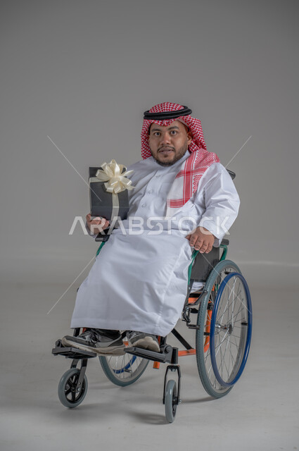 دعم المجتمع السعودي لذوي الهمم, مناسبات و أعياد سعيدة، بورتريه لرجل عربي خليجي سعودي مقعد يرتدي الثوب التقليدي والشماغ يجلس على كرسي متحرك يحمل بيده صندوق هدايا ، شعور المعاقين بالحب و التقدير، صورة لكامل الجسم، خلفية رمادية