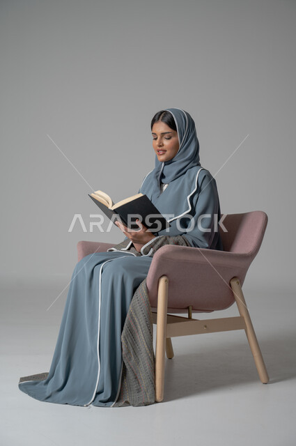 Spending time reading, learning and educating yourself, portrait of a veiled Saudi Arabian Gulf woman wearing an abaya sitting on a comfortable chair and holding a book in her hands, enjoying reading books, stories and novels, self-learning culture, colorful background
