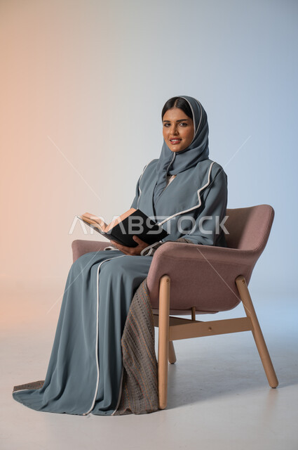 Spending time reading, learning and educating yourself, portrait of a veiled Saudi Arabian Gulf woman wearing an abaya sitting on a comfortable chair and holding a book in her hands, enjoying reading books, stories and novels, self-learning culture, colorful background