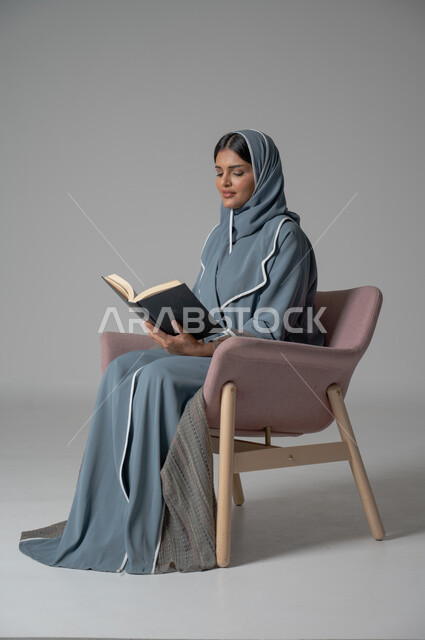 Spending time reading, learning and educating yourself, portrait of a veiled Saudi Arabian Gulf woman wearing an abaya sitting on a comfortable chair and holding a book in her hands, enjoying reading books, stories and novels, self-learning culture, colorful background