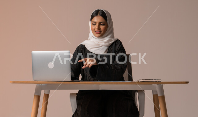 Performing tasks for professional development and achieving goals, managing and organizing business affairs, using modern and advanced technologies, portrait of a veiled Saudi Arabian Gulf woman, smiling, sitting on the chair pointing with her index finger to the laptop, beige background