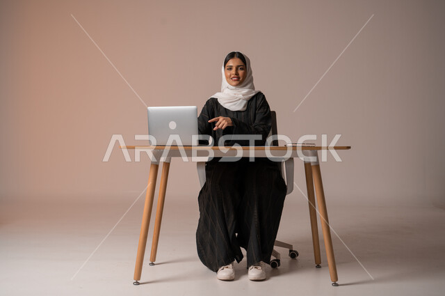 إدارة شؤون الأعمال وتنظيمها، استخدام التقنيات الحديثة والمتطورة، بورتريه لامراة عربية خليجية سعودية محجبة مبتسمة تنظر الى الكميرا تجلس على الكرسي تشير بإصبع السبابة إلى الحاسوب المحمول، اداء المهام للتطور المهني وتحقيق الاهداف, خلفية بيج
