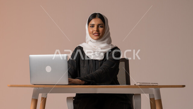 إدارة شؤون الأعمال وتنظيمه، ايماءات السعادة والسرور، استخدام التقنيات الحديثة والمتطورة، بورتريه لامراة عربية خليجية سعودية محجبة مبتسمة تنظر الى الكميرا تجلس على الكرسي أمام الحاسوب المحمول، خلفية بيج 