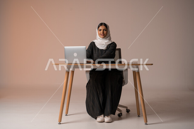 Managing and organizing business affairs, gestures of happiness and pleasure, using modern and advanced technologies, portrait of a veiled Saudi Gulf Arab woman, smiling, looking at the camera, sitting on the chair in front of the laptop, beige background