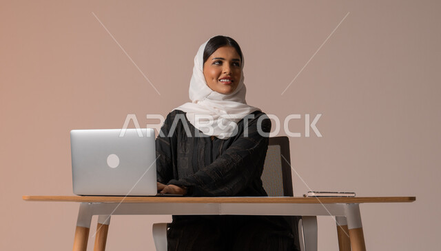 استخدام التقنيات الحديثة والمتطورة، بورتريه لامراة عربية خليجية سعودية محجبة مبتسمة تنظر للاعلى تجلس على الكرسي أمام الحاسوب المحمول، إدارة شؤون الأعمال وتنظيمه، ايماءات السعادة والسرور، خلفية بيج 
