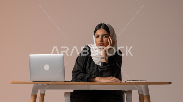 الشعور بالضجر والملل, استخدام التقنيات الحديثة المتطورة، بورتريه لامراة عربية خليجية سعودية محجبة ترتدي العباءة السوداء تجلس على الكرسي تضع يدها على ذقنها بإيماءات الملل, محاولة ايجاد حل لمشكلة ما, شرود الذهن والتفكير, خلفية بيج