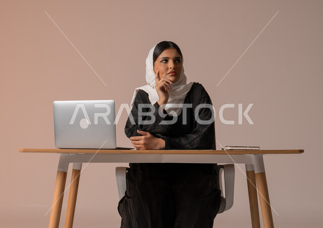 إيماءات تدل على التركيز والحيرة، التمعن و التفكير بشيء ما , التخطيط للمستقبل و إيجاد حلول للمشاكل , بورتريه لامراة عربية خليجية سعودية محجبة ترتدي العباءة السوداء تجلس على مكتبها أمام الحاسوب المحمول، استخدام التقنيات الحديثة المتطورة, خلفية بيج