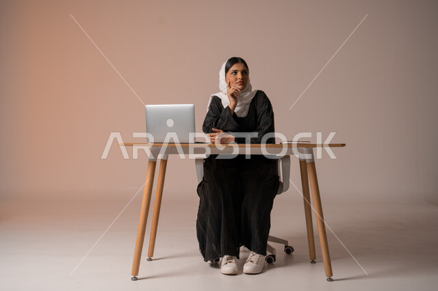 إيماءات تدل على التركيز والحيرة، التمعن و التفكير بشيء ما , التخطيط للمستقبل و إيجاد حلول للمشاكل , بورتريه لامراة عربية خليجية سعودية محجبة ترتدي العباءة السوداء تجلس على مكتبها أمام الحاسوب المحمول، استخدام التقنيات الحديثة المتطورة, خلفية بيج