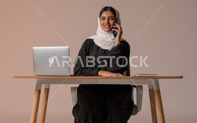 استخدام التقنيات الحديثة والمتطورة، بورتريه لامراة عربية خليجية سعودية محجبة مبتسمة ترتدي العباءة السوداء تجلس على الكرسي أمام الحاسوب المحمول مبتسمة تقوم بإجراء مكالمة صوتية عبر الجوال، تعابير الفرح عند سماع الاخبار السارة، النظر الى الكاميرا بايماءات السعادة والسرور، خلفية بيج