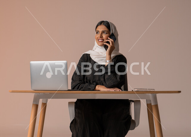 استخدام التقنيات الحديثة والمتطورة، بورتريه لامراة عربية خليجية سعودية محجبة مبتسمة ترتدي العباءة السوداء تجلس على الكرسي أمام الحاسوب المحمول مبتسمة تقوم بإجراء مكالمة صوتية عبر الجوال، تعابير الفرح عند سماع الاخبار السارة، خلفية بيج