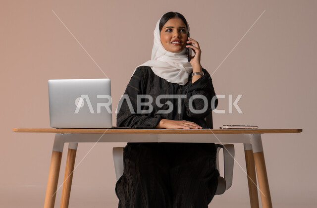 Using modern and advanced technologies, portrait of a smiling veiled Saudi Arabian Gulf woman wearing a black abaya sitting on a chair in front of a laptop smiling making a voice call via mobile, expressions of joy upon hearing good news, beige background