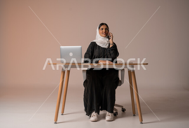 Using modern and advanced technologies, portrait of a smiling veiled Saudi Arabian Gulf woman wearing a black abaya sitting on a chair in front of a laptop smiling making a voice call via mobile, expressions of joy upon hearing good news, beige background