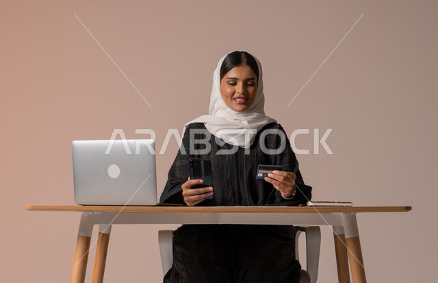 استخدام اجهزة تقنية حديثة للدفع عن بعد عبر الانترنت، إمكانية الشراء والدفع من خلال البرامج والتطبيقات الالكترونية، بورتريه لامراة عربية خليجية سعودية محجبة ترتدي عباءة تجلس على كرسي مريح أمام الحاسوب المحمول تحمل بيدها الجوال وبطاقة الائتمان البنكية للتسوق الالكتروني، خلفية بيج