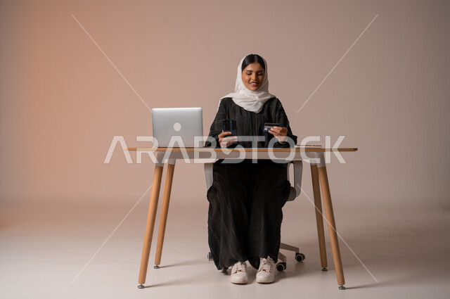 The possibility of purchasing and paying through electronic programs and applications, using modern technical devices for remote payment via the Internet, a portrait of a veiled Saudi Arabian Gulf woman wearing an abaya sitting on a comfortable chair in front of a laptop holding a mobile phone and a bank credit card for electronic shopping, beige background