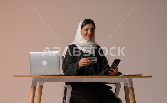 The possibility of purchasing and paying through electronic programs and applications, using modern technical devices for remote payment via the Internet, a portrait of a veiled Saudi Arabian Gulf woman wearing an abaya sitting on a comfortable chair in front of a laptop holding a mobile phone and a bank credit card for electronic shopping, beige background