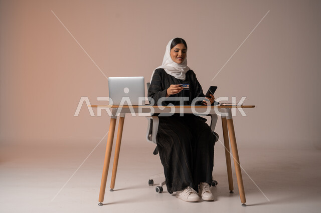 إمكانية الشراء والدفع من خلال البرامج والتطبيقات الالكترونية، استخدام اجهزة تقنية حديثة للدفع عن بعد عبر الانترنت، بورتريه لامراة عربية خليجية سعودية محجبة ترتدي عباءة تجلس على كرسي مريح أمام الحاسوب المحمول تحمل بيدها الجوال وبطاقة الائتمان البنكية للتسوق الالكتروني، خلفية بيج