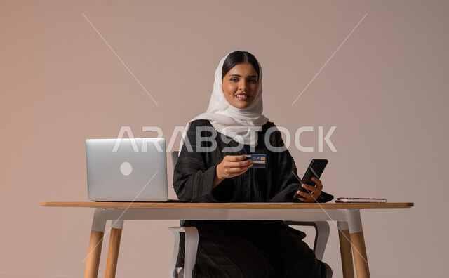 The possibility of purchasing and paying through electronic programs and applications, using modern technical devices for remote payment via the Internet, a portrait of a veiled Saudi Arabian Gulf woman wearing an abaya sitting on a comfortable chair in front of a laptop holding a mobile phone and a bank credit card for electronic shopping, beige background