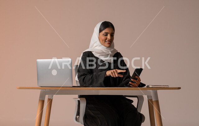 Using modern and advanced technologies, portrait of a smiling veiled Saudi Arabian Gulf woman wearing a black abaya sitting on a chair in front of a laptop and holding a mobile phone in her hand, communicating with family and friends remotely, managing and organizing business affairs, beige background