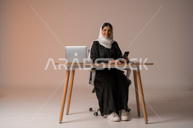 Using modern and advanced technologies, a portrait of a smiling, veiled Saudi Arabian Gulf woman wearing a black abaya, sitting on the chair in front of the laptop and holding the mobile phone in her hand, communicating with family and friends remotely, managing and organizing business affairs, looking at the camera with gestures of happiness and pleasure, beige background.