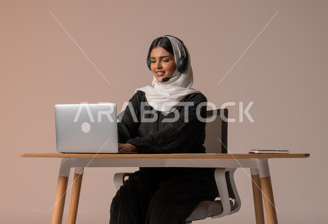 استخدام التقنيات الحديثة والمتطورة، الرد على الاستعلامات والاستفسارات وخدمة العملاء، بورتريه مقرب لامراة عربية خليجية سعودية محجبة مبتسمة ترتدي العباءة السوداء تجلس على الكرسي أمام الحاسوب المحمول وتضع سماعة الهيدفون، التواصل مع الأهل و الأصدقاء عن بعد، خلفية بيج