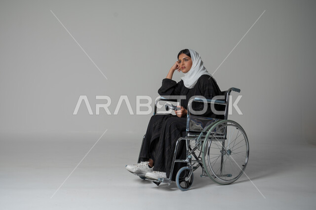Absent-mindedness on a specific topic, gestures of contemplation and reflection, expressions of integration and confusion, portrait of a Saudi Arabian Gulf woman with special needs sitting in a wheelchair, wearing a hijab, putting her hand on her cheek and thinking, finding solutions to a problem, gray background