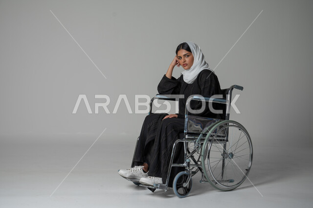 شرود الذهن في موضوع معين، إيماءات التأمل والتمعن, تعابير الاندماج و الحيرة , بورتريه لإمرأة عربية سعودية خليجية من ذوي الاحتياجات الخاصه تجلس على كرسي متحرك محجبة تضع يدها خدها وتفكر, إيجاد الحلول لمشكلة ما، خلفية رمادية