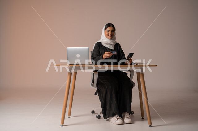 إمكانية الشراء والدفع من خلال البرامج والتطبيقات الالكترونية، استخدام اجهزة تقنية حديثة للدفع عن بعد عبر الانترنت، بورتريه لامراة عربية خليجية سعودية محجبة ترتدي عباءة تجلس على كرسي مريح أمام الحاسوب المحمول تحمل بيدها الجوال وبطاقة الائتمان البنكية للتسوق الالكتروني، خلفية بيج
