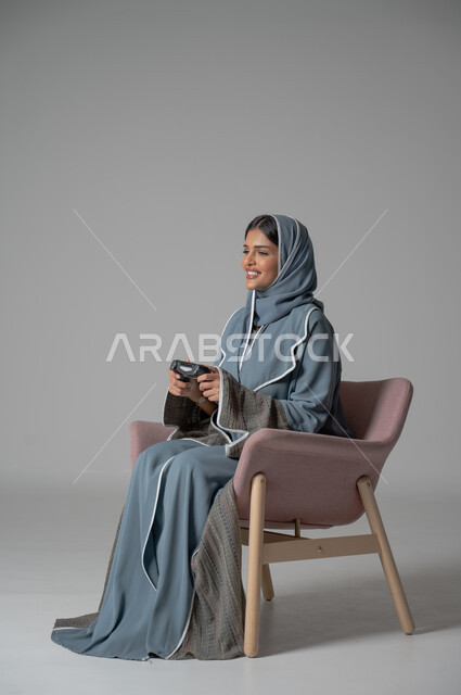 قضاء أوقات الفراغ بشكل ممتع، استخدام التقنيات الحديثة, بورتريه لامرأة عربية خليجية سعودية محجبة ترتدي العباءة تجلس على كرسي مريح تحمل بيدها جهاز التحكم بايماءات السعادة والسرور أثناء اللعب, خلفية رمادية