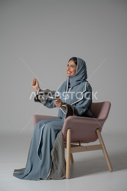 استخدام التقنيات الحديثة, قضاء أوقات الفراغ بشكل ممتع، بورتريه لامرأة عربية خليجية سعودية محجبة ترتدي العباءة تجلس على كرسي مريح تحمل بيدها جهاز التحكم بايماءات التفاعل والاندماج أثناء اللعب, خلفية رمادية