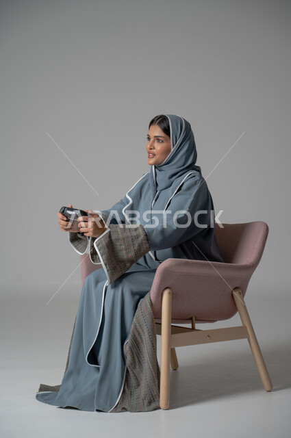 قضاء أوقات الفراغ بشكل ممتع، استخدام التقنيات الحديثة, بورتريه لامرأة عربية خليجية سعودية محجبة ترتدي العباءة تجلس على كرسي مريح تحمل بيدها جهاز التحكم بايماءات السعادة والسرور أثناء اللعب, خلفية رمادية