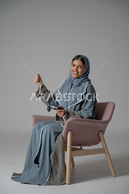 استخدام التقنيات الحديثة, قضاء أوقات الفراغ بشكل ممتع، بورتريه لامرأة عربية خليجية سعودية محجبة ترتدي العباءة تجلس على كرسي مريح تحمل بيدها جهاز التحكم بايماءات التفاعل والاندماج أثناء اللعب, خلفية رمادية