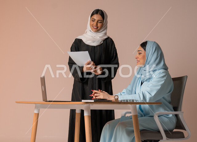 الشراكة والتعاون في إنجاز المهام، بورتريه لموظفة عربية خليجية سعودية محجبة ترتدي العباءة السوداء تحمل بيدها اوراق العمل تقوم بالتأكد من صحة المعلومات من سيدة الاعمال، دراسة وتحليل المشاريع الجديدة، اداء المهام للتطور المهني وتحقيق الاهداف, خلفية بيج