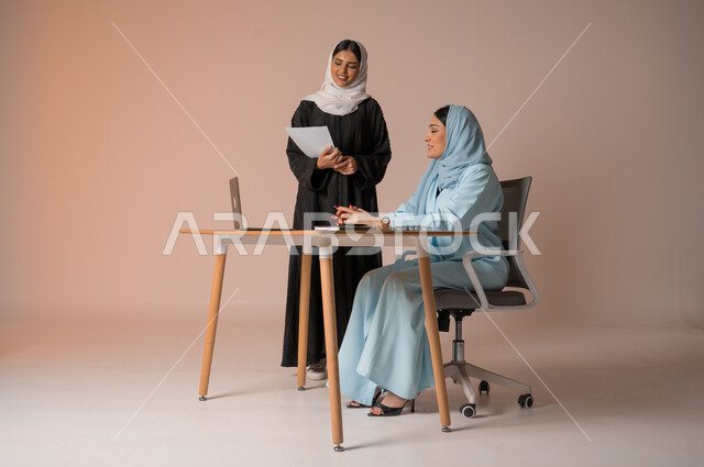 الشراكة والتعاون في إنجاز المهام، بورتريه لموظفة عربية خليجية سعودية محجبة ترتدي العباءة السوداء تحمل بيدها اوراق العمل تقوم بالتأكد من صحة المعلومات من سيدة الاعمال، دراسة وتحليل المشاريع الجديدة، اداء المهام للتطور المهني وتحقيق الاهداف, خلفية بيج