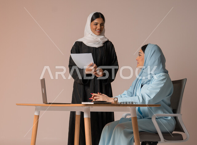 الشراكة والتعاون في إنجاز المهام، بورتريه لموظفة عربية خليجية سعودية محجبة ترتدي العباءة السوداء تحمل بيدها اوراق العمل تقوم بالتأكد من صحة المعلومات من سيدة الاعمال، دراسة وتحليل المشاريع الجديدة، اداء المهام للتطور المهني وتحقيق الاهداف, خلفية بيج