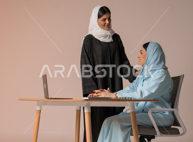 الشراكة والتعاون في إنجاز المهام، بورتريه لسيدتان عربيتان خليجيتات سعوديتان محجبتان في اجتماع عمل، استخدام جهاز الحاسوب المحمول لمتابعة ومناقشة الأعمال، دراسة وتحليل المشاريع الجديدة، اداء المهام للتطور المهني وتحقيق الاهداف, خلفية بيج