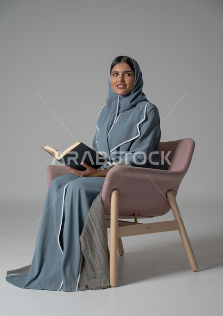 قضاء الوقت بالقراءة والتعلم والتثقف، بورتريه لامرأة عربية سعودية خليجية محجبة ترتدي العباءة تجلس على كرسي مريح وتحمل بيديها كتاب، الاستمتاع بقراءة الكتب والقصص والروايات, ثقافة التعلم الذاتي, خلفية رمادية