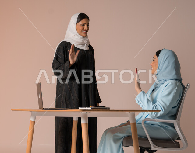 تبادل الخبرات والاتفاقيات الناجحة في مكتب الشركه، مفهوم الإدارة المتعاونة بين الموظفين، بورتريه لصديقتين عربيتين خليجيتين سعوديتين مبتسمتين ترتديان العباءة يقومان بالتلويح باليد و السلام عن بعد، استخدام جهاز الحاسوب المحمول لمتابعة ومناقشة الأعمال, خلفية بيج