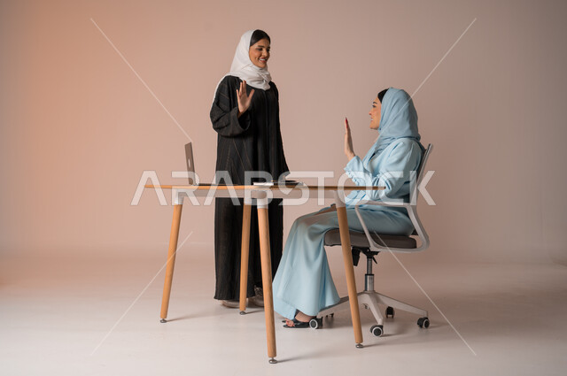 تبادل الخبرات والاتفاقيات الناجحة في مكتب الشركه، مفهوم الإدارة المتعاونة بين الموظفين، بورتريه لصديقتين عربيتين خليجيتين سعوديتين مبتسمتين ترتديان العباءة يقومان بالتلويح باليد و السلام عن بعد، استخدام جهاز الحاسوب المحمول لمتابعة ومناقشة الأعمال, خلفية بيج