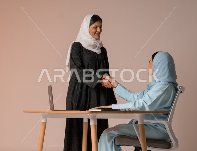 عقد صفقات شراكة جديدة مع أطراف متنوعة لتوسيع مجال العمل، مفهوم الإدارة المتعاونة، بورتريه لأمرأتين عربيتين سعوديتين خليجيتين محجبتين مبتسمتين ترتديان العباءة تتصافحان بإيماءات السرور لنجاح خطة الشركة، التعاون وتحقيق مكاسب وأرباح عالية، خلفية بيج