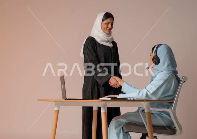 عقد صفقات شراكة جديدة مع أطراف متنوعة لتوسيع مجال العمل، مفهوم الإدارة المتعاونة، بورتريه لأمرأتين عربيتين سعوديتين خليجيتين محجبتين مبتسمتين ترتديان العباءة تتصافحان بإيماءات السرور لنجاح خطة الشركة، التعاون وتحقيق مكاسب وأرباح عالية، خلفية بيج