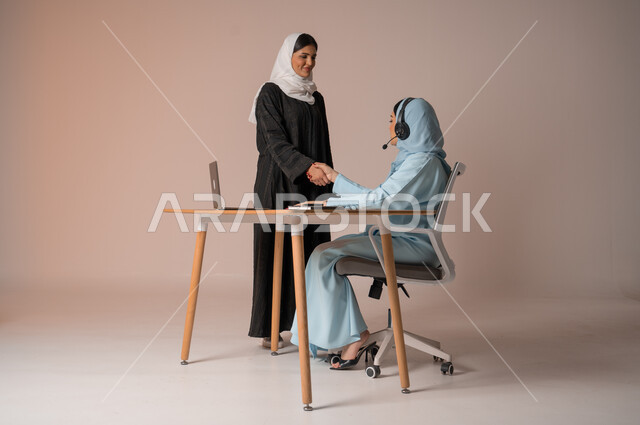 عقد صفقات شراكة جديدة مع أطراف متنوعة لتوسيع مجال العمل، مفهوم الإدارة المتعاونة، بورتريه لأمرأتين عربيتين سعوديتين خليجيتين محجبتين مبتسمتين ترتديان العباءة تتصافحان بإيماءات السرور لنجاح خطة الشركة، التعاون وتحقيق مكاسب وأرباح عالية، خلفية بيج
