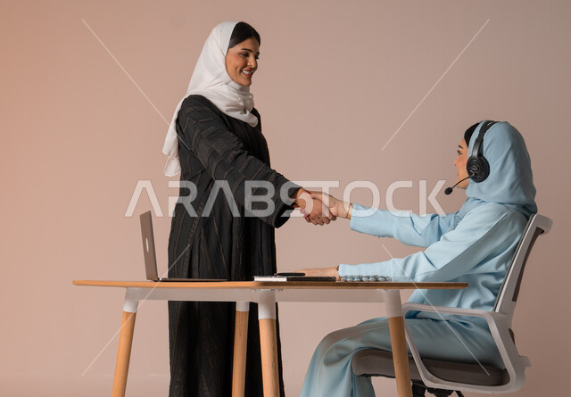 عقد صفقات شراكة جديدة مع أطراف متنوعة لتوسيع مجال العمل، مفهوم الإدارة المتعاونة، بورتريه لأمرأتين عربيتين سعوديتين خليجيتين محجبتين مبتسمتين ترتديان العباءة تتصافحان بإيماءات السرور لنجاح خطة الشركة، التعاون وتحقيق مكاسب وأرباح عالية، خلفية بيج