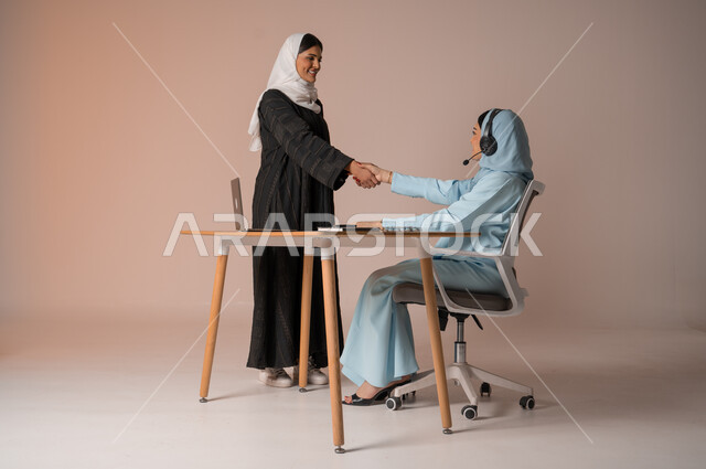 عقد صفقات شراكة جديدة مع أطراف متنوعة لتوسيع مجال العمل، مفهوم الإدارة المتعاونة، بورتريه لأمرأتين عربيتين سعوديتين خليجيتين محجبتين مبتسمتين ترتديان العباءة تتصافحان بإيماءات السرور لنجاح خطة الشركة، التعاون وتحقيق مكاسب وأرباح عالية، خلفية بيج