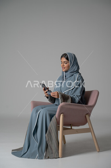 التواصل مع الأهل و الأصدقاء عن بعد, استخدام الاجهزة الحديثة والتقنية, دمج التكنولوجيا المتطورة في الحياة اليومية، بورتريه لامرأة أنيقة عربية خليجية سعودية محجبة ترتدي العباءة تجلس على كرسي مريح تحمل الهاتف المحمول بإيماءات السعادة والسرور, خلفية رمادية