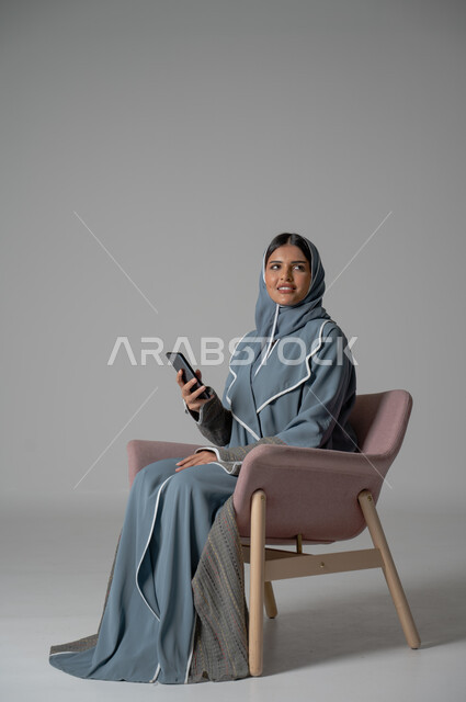 التواصل مع الأهل و الأصدقاء عن بعد, استخدام الاجهزة الحديثة والتقنية, دمج التكنولوجيا المتطورة في الحياة اليومية، بورتريه لامرأة أنيقة عربية خليجية سعودية محجبة ترتدي العباءة تجلس على كرسي مريح تحمل الهاتف المحمول بإيماءات السعادة والسرور, خلفية رمادية
