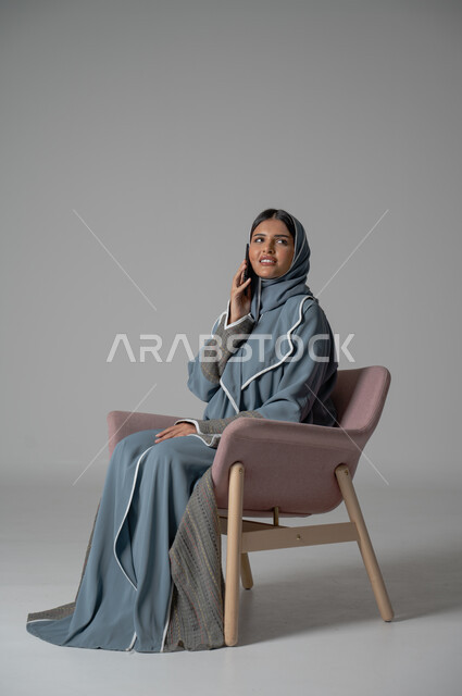 التواصل مع الأهل و الأصدقاء عن بعد, استخدام الاجهزة الحديثة والتقنية, دمج التكنولوجيا المتطورة في الحياة اليومية، بورتريه لامرأة أنيقة عربية خليجية سعودية محجبة ترتدي العباءة تجلس على كرسي مريح تحمل الهاتف المحمول بإيماءات السعادة والسرور, خلفية رمادية