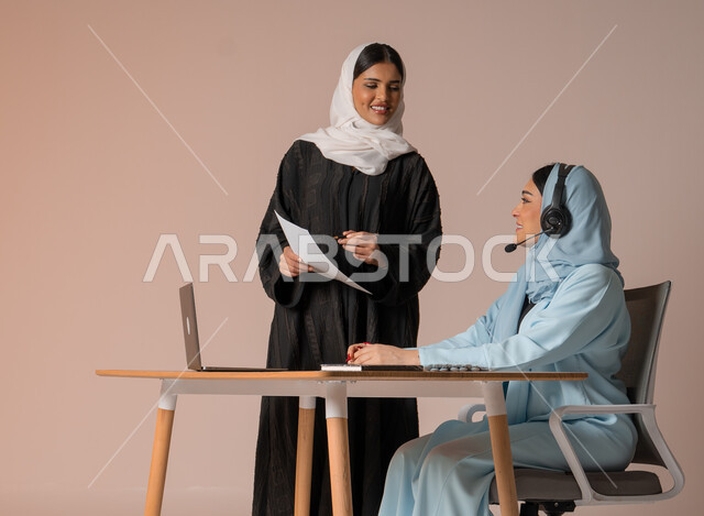 Study and analysis of new projects, performing tasks for professional development and achieving goals, partnership and cooperation in completing tasks, portrait of a veiled Saudi Gulf Arab employee wearing a black abaya, holding work papers in her hand, verifying the accuracy of information from the businesswoman, beige background