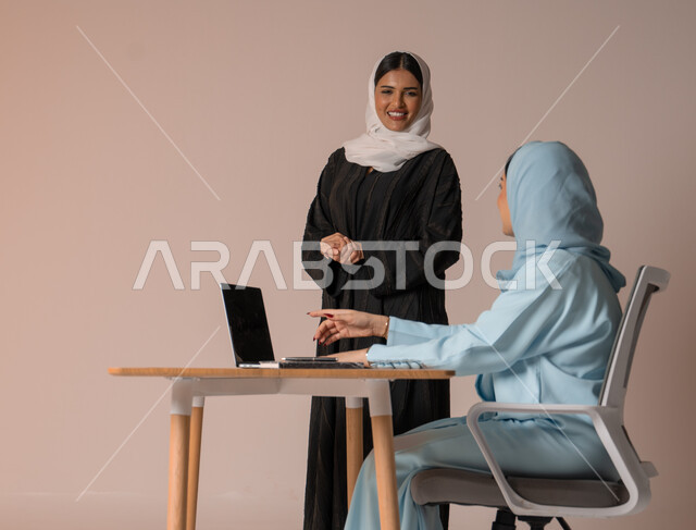  استخدام جهاز الحاسوب المحمول لمتابعة ومناقشة الأعمال، اداء المهام للتطور المهني وتحقيق الاهداف, الشراكة والتعاون في إنجاز المهام، بورتريه لسيدتان عربيتان خليجيتات سعوديتان محجبتان في اجتماع عمل، دراسة وتحليل المشاريع الجديدة، خلفية بيج
