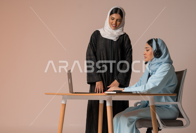 Performing tasks for professional development and achieving goals, partnership and cooperation in completing tasks, a portrait of two veiled Saudi Gulf Arab women in a business meeting, using the laptop to follow up and discuss work, studying and analyzing new projects, beige background