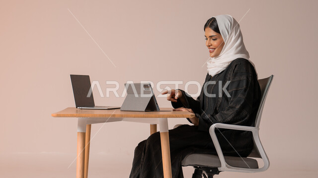 استخدام التقنيات الحديثة والمتطورة، بورتريه لامراة عربية خليجية سعودية محجبة مبتسمة ترتدي العباءة السوداء تجلس على الكرسي أمام الحاسوب المحمول مبتسمة تشير بأصبع السبابة الى التابليت تقوم بإجراء مكالمة فيديو، التواصل مع الأهل و الأصدقاء عن بعد، خلفية بيج